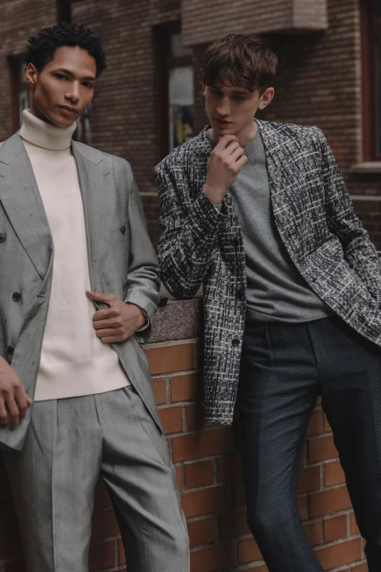 Two adult men in stylish blazers and dress pants pose against a brick wall in an urban setting, showcasing contemporary fashion.