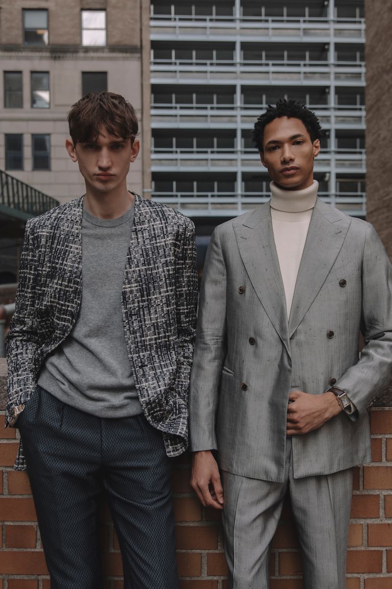 Two men model tailored suits against a brick wall in a city setting, showcasing modern fashion and urban style in natural lighting.