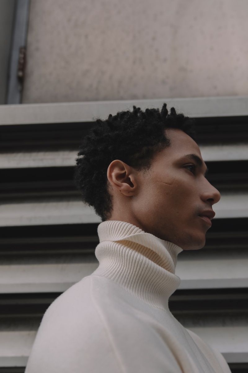 Portrait of a young adult man with a neutral expression wearing a turtleneck sweater, posing in front of a gray textured background.