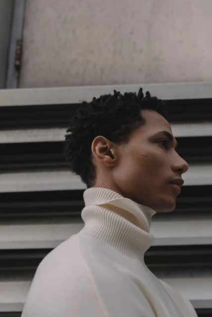 Portrait of a young adult man with a neutral expression wearing a turtleneck sweater, posing in front of a gray textured background.