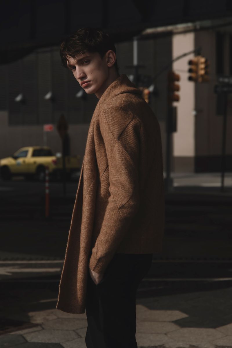 Young man in brown coat stands on a city street with traffic light in background, creating a stylish outdoor fashion portrait.