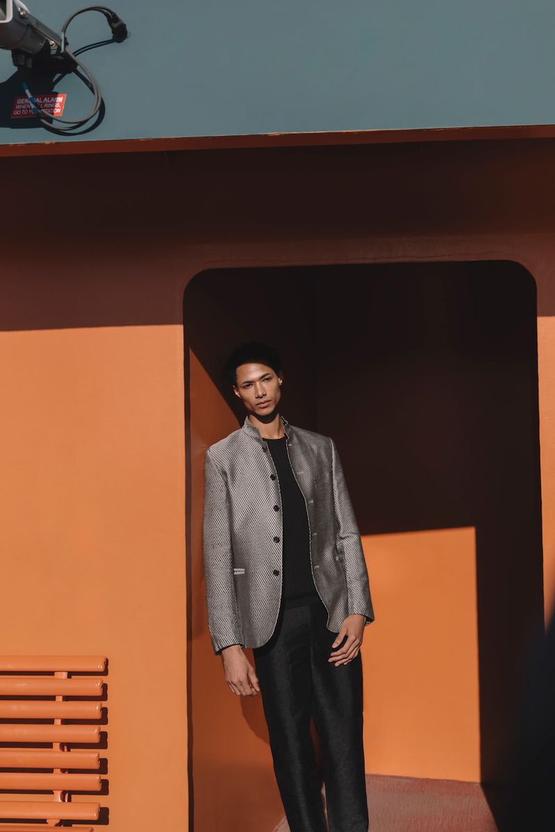 A stylish adult male model is posing outdoors in a textured blazer, a black shirt and black pants, in front of an orange wall.