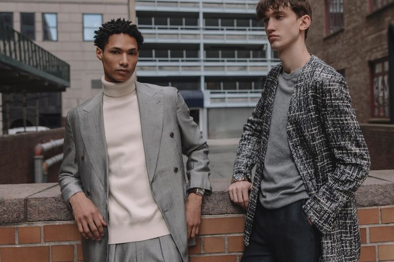 Two adult men in stylish outerwear, standing on a city rooftop with brick walls and a tall building in the background, fashion shoot.