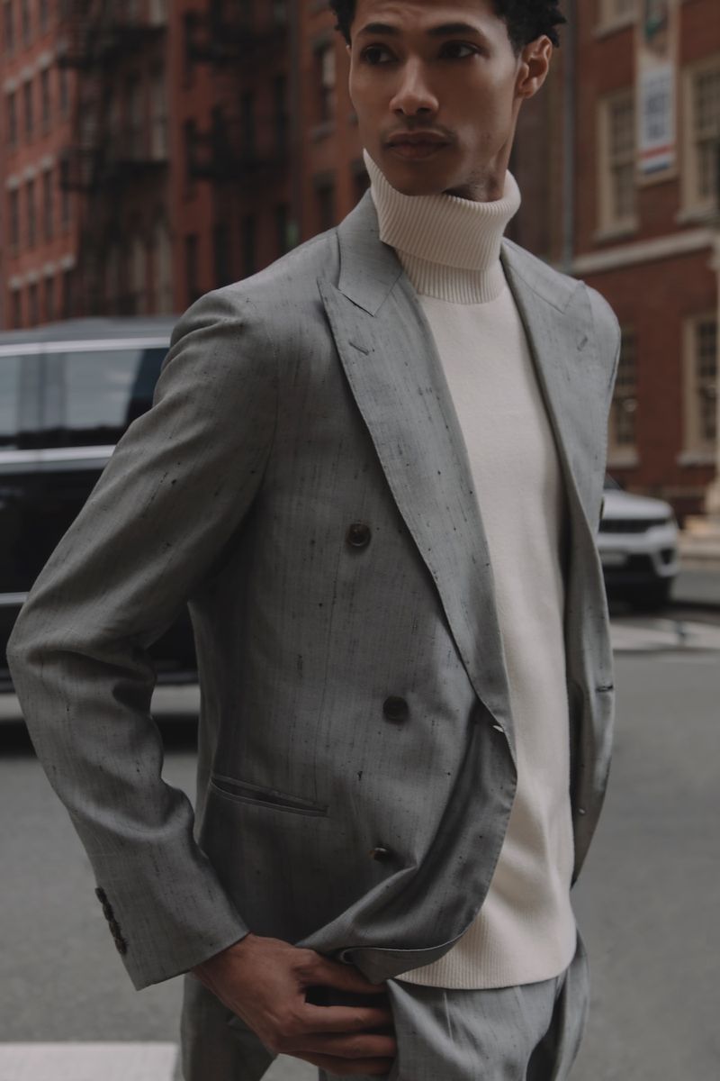 A well-dressed man in a tailored suit poses on a city street, with classic brick buildings and parked cars forming the urban backdrop.