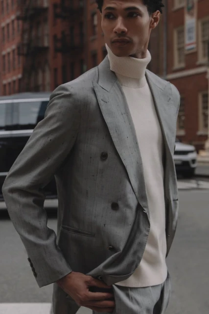 A well-dressed man in a tailored suit poses on a city street, with classic brick buildings and parked cars forming the urban backdrop.