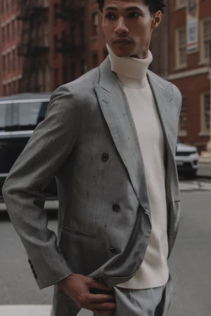 A well-dressed man in a tailored suit poses on a city street, with classic brick buildings and parked cars forming the urban backdrop.