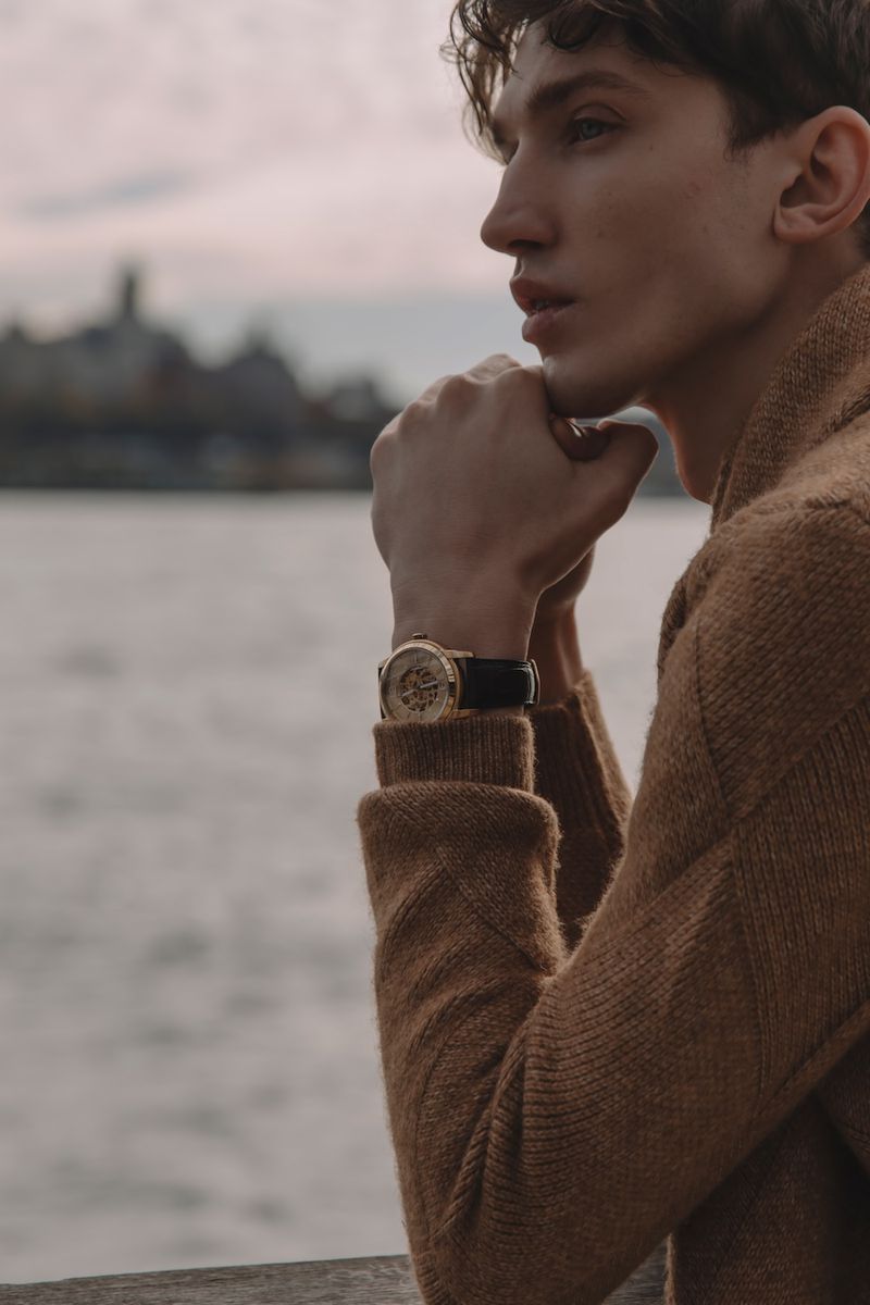 Adult male poses wearing a brown knit sweater and a wristwatch. He is outdoors with a blurred cityscape and body of water.