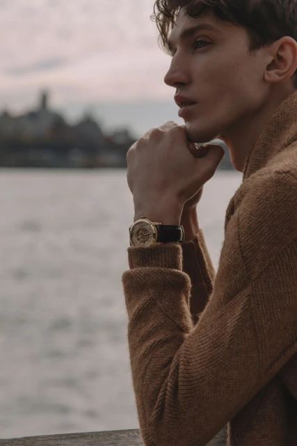 Adult male poses wearing a brown knit sweater and a wristwatch. He is outdoors with a blurred cityscape and body of water.