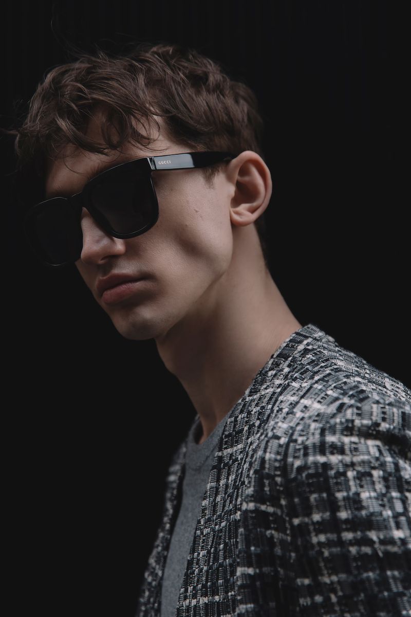 Man with dark hair and tweed jacket wearing black sunglasses in a studio setting with a dark background and diffused lighting.
