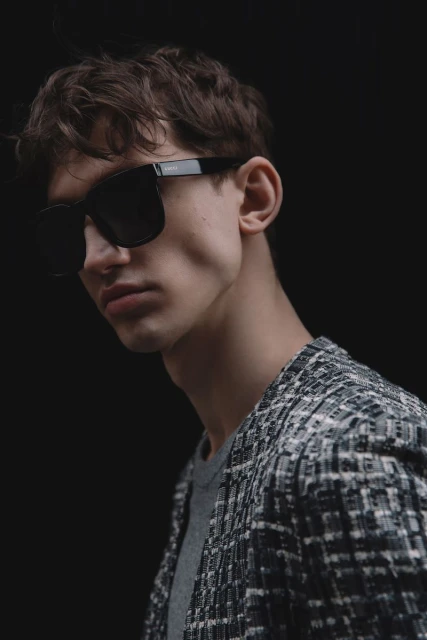 Man with dark hair and tweed jacket wearing black sunglasses in a studio setting with a dark background and diffused lighting.
