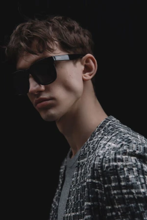 Man with dark hair and tweed jacket wearing black sunglasses in a studio setting with a dark background and diffused lighting.