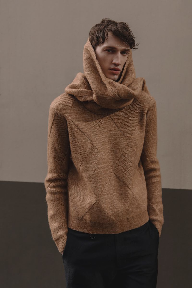 Portrait of a male fashion model in a brown sweater with a scarf-like hood, standing against a neutral background in soft, diffused lighting.