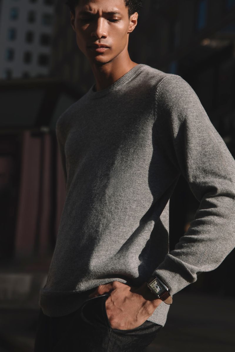 An adult man in a grey sweater poses in an outdoor setting with buildings in the background, bathed in natural sunlight, wearing a watch.