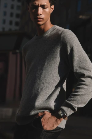 An adult man in a grey sweater poses in an outdoor setting with buildings in the background, bathed in natural sunlight, wearing a watch.