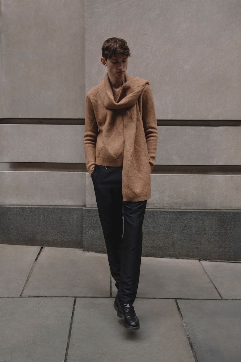 A young adult male walking in a neutral-toned sweater, scarf, and dark pants, outside against a concrete wall with horizontal lines.