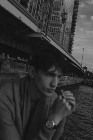 Black and white portrait of a young adult male in an urban setting with skyscrapers and a waterfront background, wearing a watch.