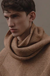 Close-up portrait of a male model wearing a thick camel-colored knit sweater with a high rolled collar against a neutral grey wall.