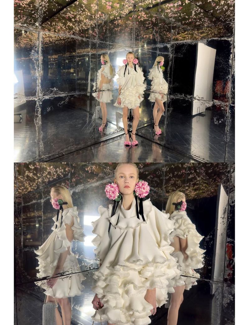 Three fashion models in matching ruffled white dresses and pink heels pose inside a mirrored room with a floral motif for a fashion shoot.