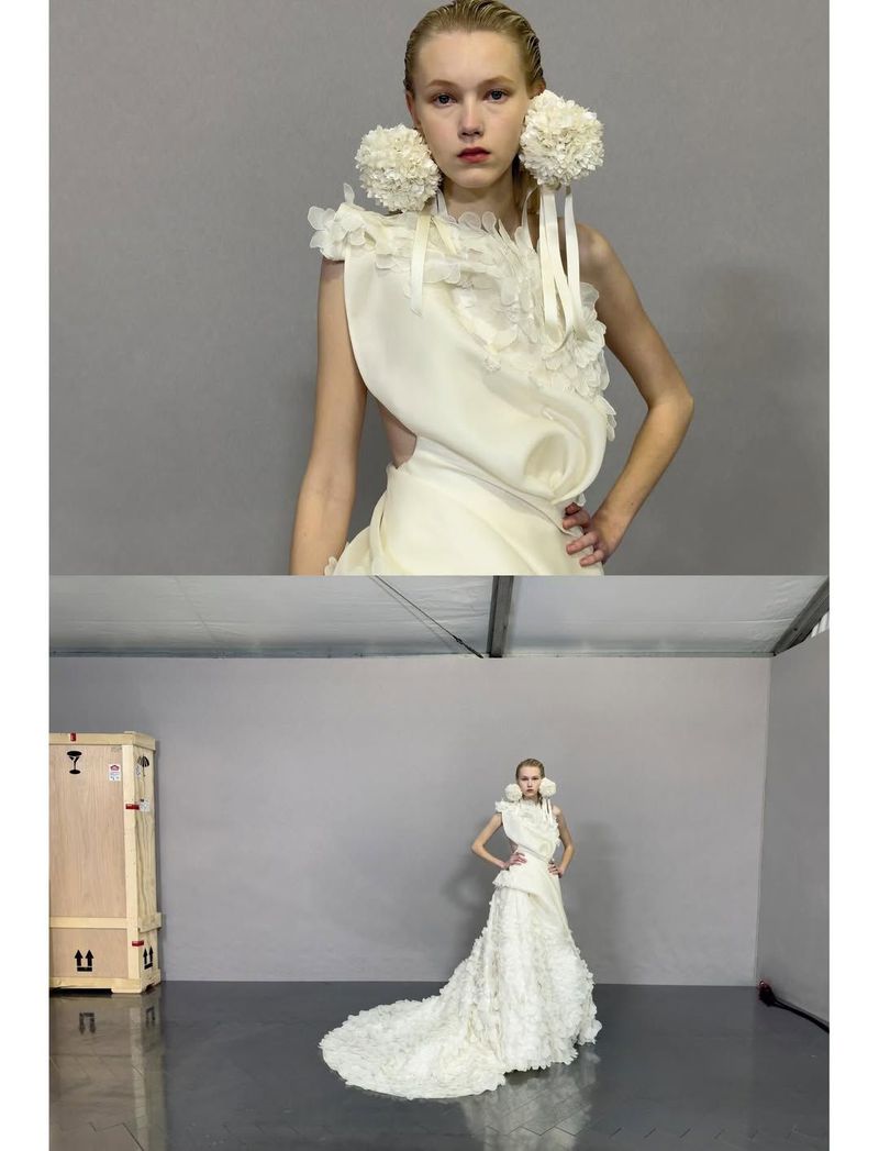 Runway model in an ivory gown with pom-pom earrings poses in a studio, captured in medium full shot with a neutral background.