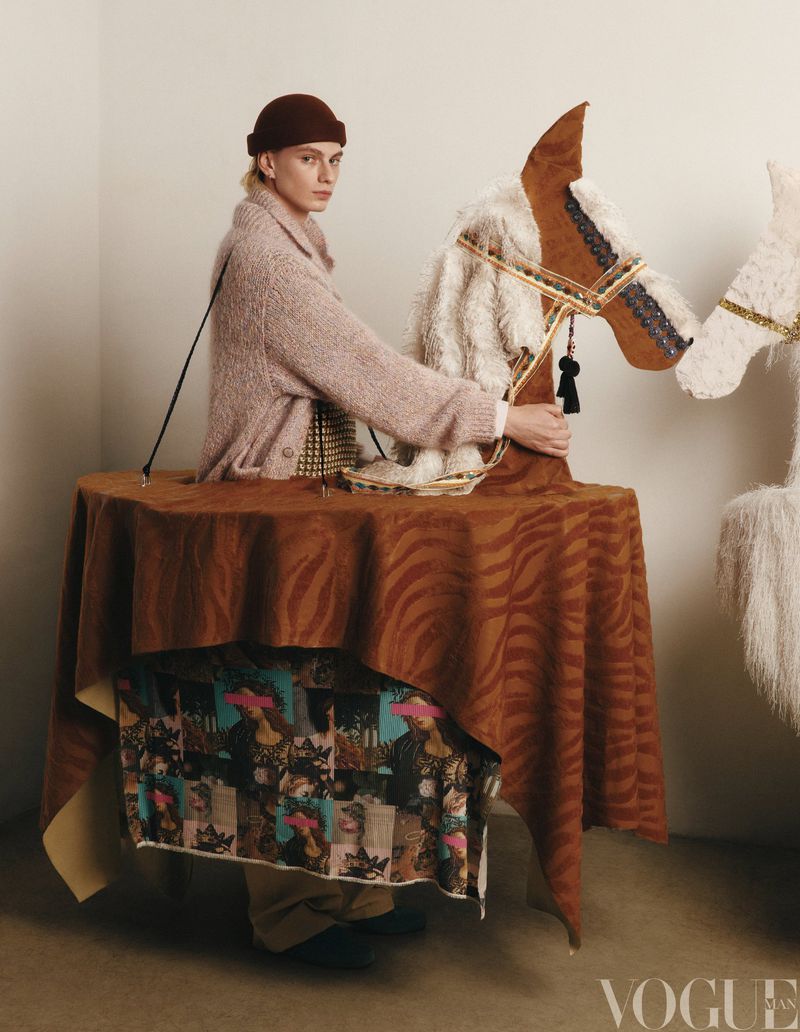 A person in a brown hat and pink sweater riding a brown hobby horse with a patterned tablecloth, fashion editorial photography.