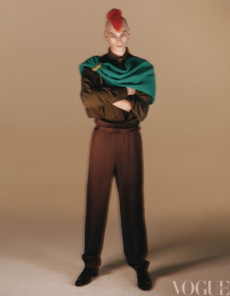 Full-length portrait of a person with a red mohawk, wearing a green shawl and brown shirt and trousers, posing indoors against a beige background.