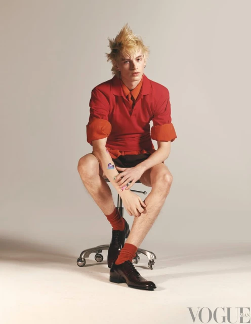 Blonde male model sits in a photo studio wearing a red shirt, black shorts, and dress shoes, styled for a fashion editorial.