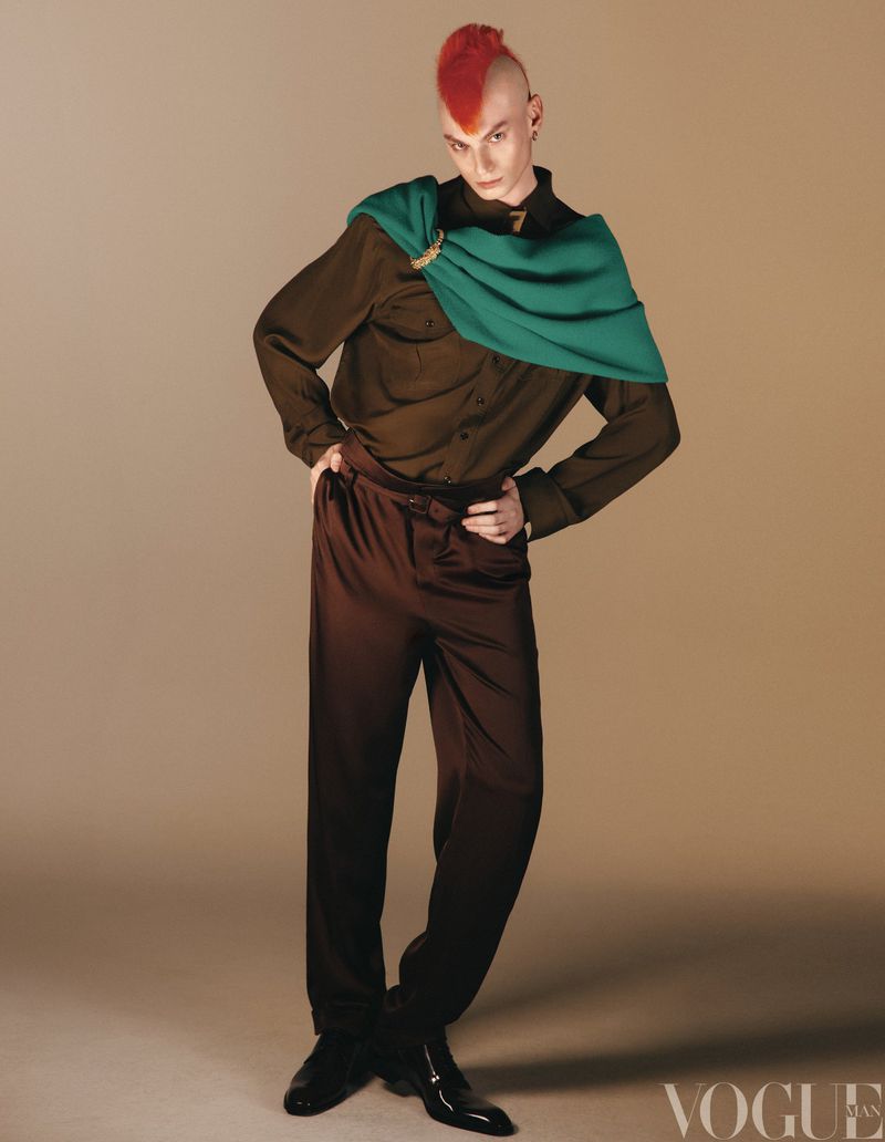 Full-length portrait of a person with a red mohawk wearing a green scarf, brown shirt, and pants against a tan background in a studio.