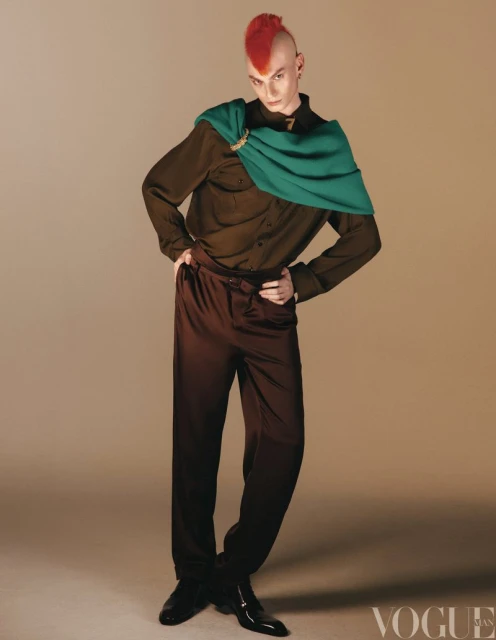 Full-length portrait of a person with a red mohawk wearing a green scarf, brown shirt, and pants against a tan background in a studio.