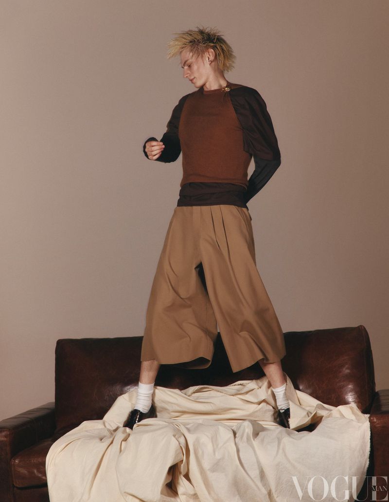 Androgynous fashion model poses atop a vintage couch in an editorial shot featuring a brown top, tan culottes, and short spiky blond hair.