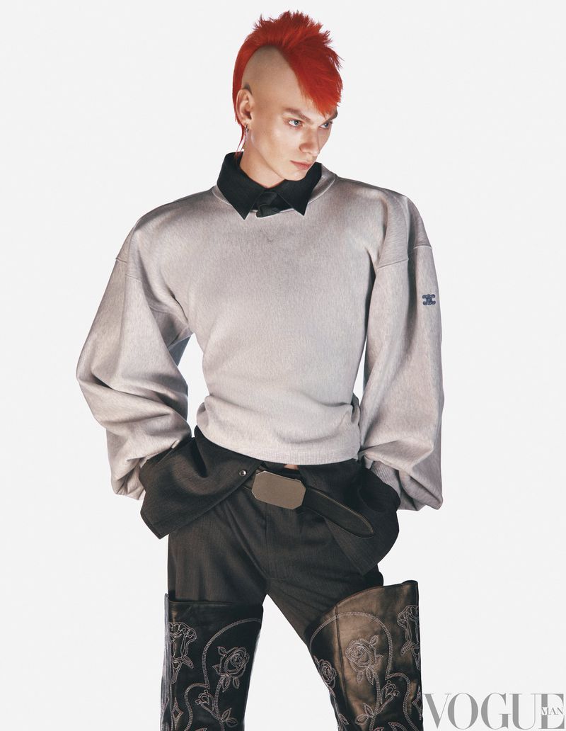Fashionable person with a vibrant red mohawk, wearing a gray sweater, black shirt and pants, and black cowboy boots against white backdrop.