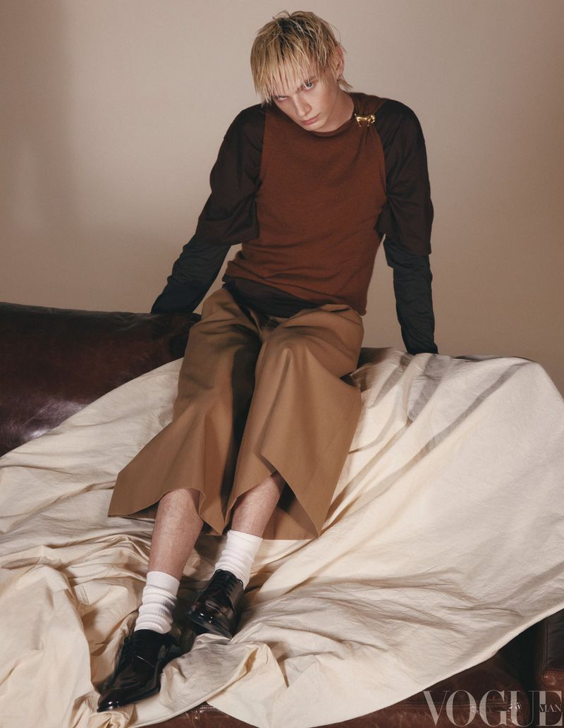 Fashion shot of a young adult with blond hair in a two-tone shirt and wide-leg pants sitting on a couch with a cream-colored fabric.
