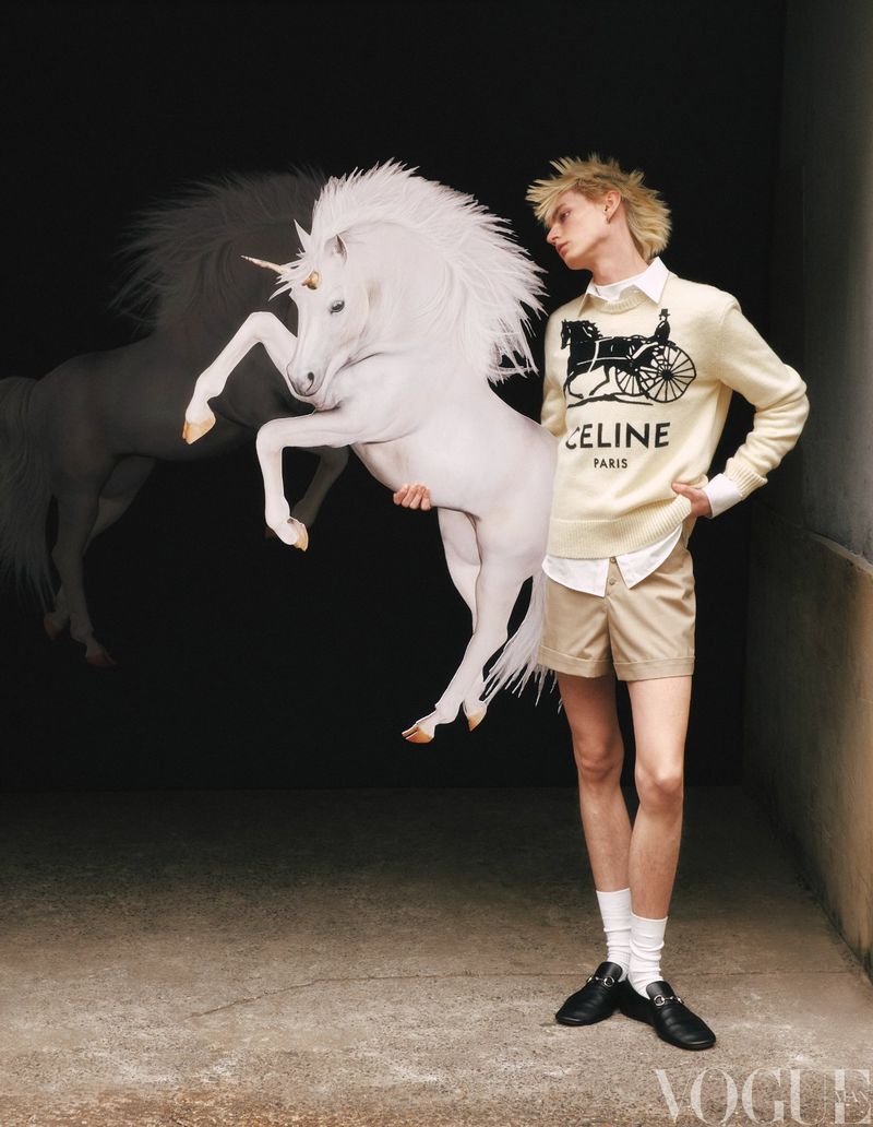 Male model poses indoors holding a white toy unicorn, wearing a cream-colored sweater, khaki shorts, and black loafers in a studio setting.