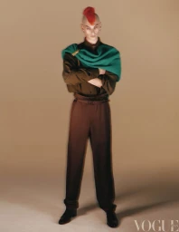 Model with red mohawk wearing a green sweater draped over a dark shirt and trousers against a neutral background.