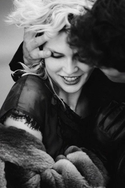 Close-up black and white shot of an adult female smiling, her hair being touched by an adult male. The shot is intimate and soft.