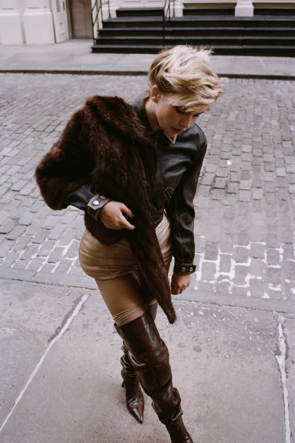 Woman in a leather jacket and thigh-high boots stands on a cobblestone street holding a fur coat, with buildings in the background.