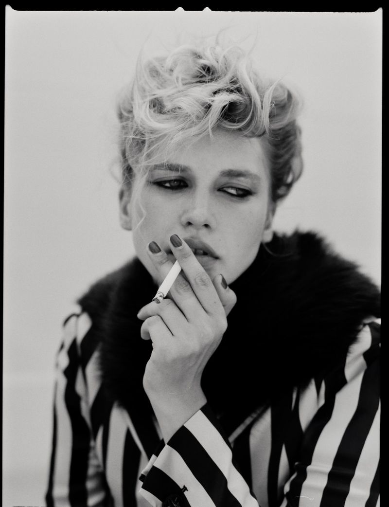 Black and white portrait of a young fair-skinned woman with wavy blonde hair, smoking a cigarette indoors, wearing a striped top.