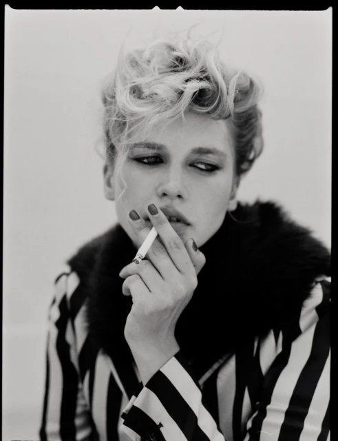 Black and white portrait of a young fair-skinned woman with wavy blonde hair, smoking a cigarette indoors, wearing a striped top.