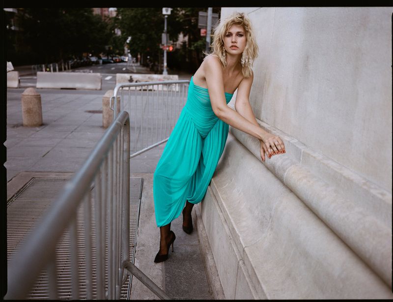 Woman in a vibrant turquoise strapless jumpsuit leaning against a stone wall on a city street.