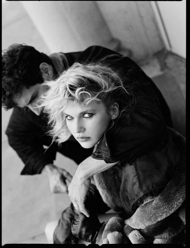 Monochrome editorial portrait of a blonde woman in dark clothing with a man behind her for Domina Journal