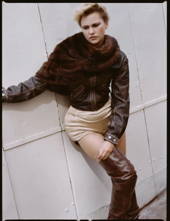 A blonde woman poses leaning against a wall in a leather jacket, fur collar, tan shorts, and brown thigh-high boots in a fashion shot.