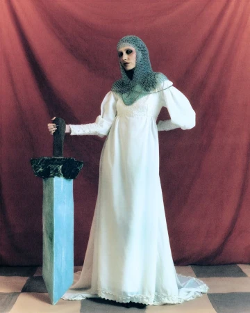 Woman in a white dress and chainmail coif poses with a large sword against a red backdrop and checkered floor. Full shot.