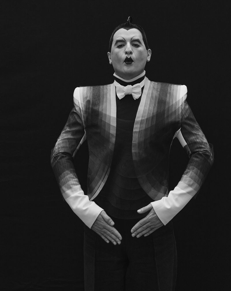 Adult male poses in a black and white photo, wearing a patterned jacket with a bow tie, against a plain black backdrop in a studio.