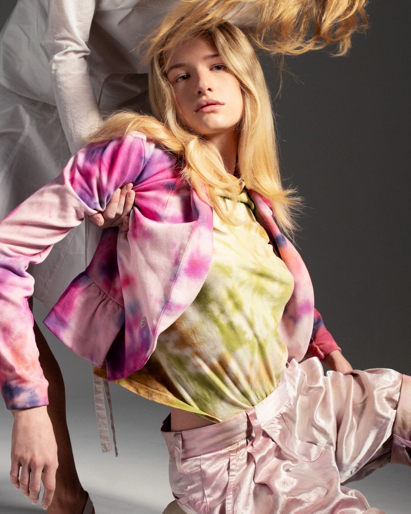 Fashion shot of a young adult female posing in a tie-dye patterned top and pink satin shorts with an off-white backdrop.