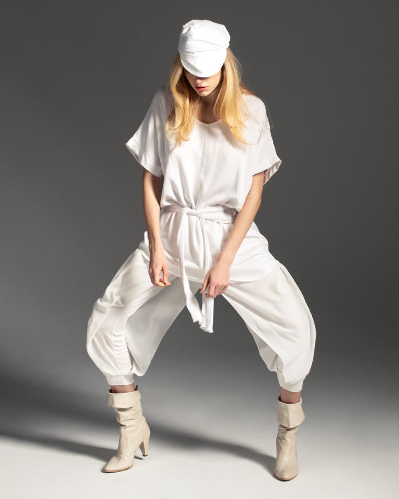 A blonde woman poses in all-white attire against a gray backdrop, featuring a loose shirt, capri pants, ankle boots, and a white cap.