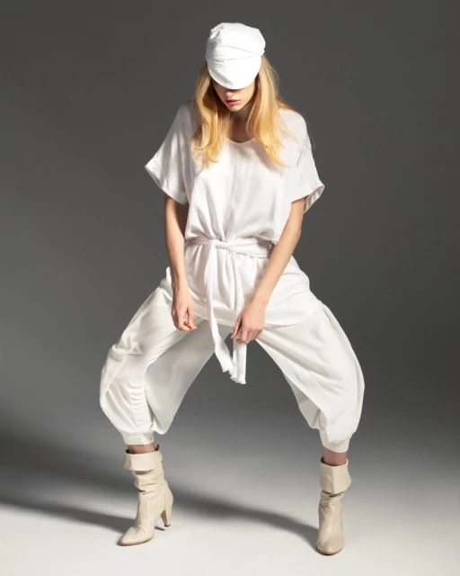 A blonde woman poses in all-white attire against a gray backdrop, featuring a loose shirt, capri pants, ankle boots, and a white cap.