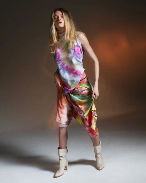 Young woman poses in tie-dye top and shorts with beige heeled boots in a studio setting with a soft, diffused light.