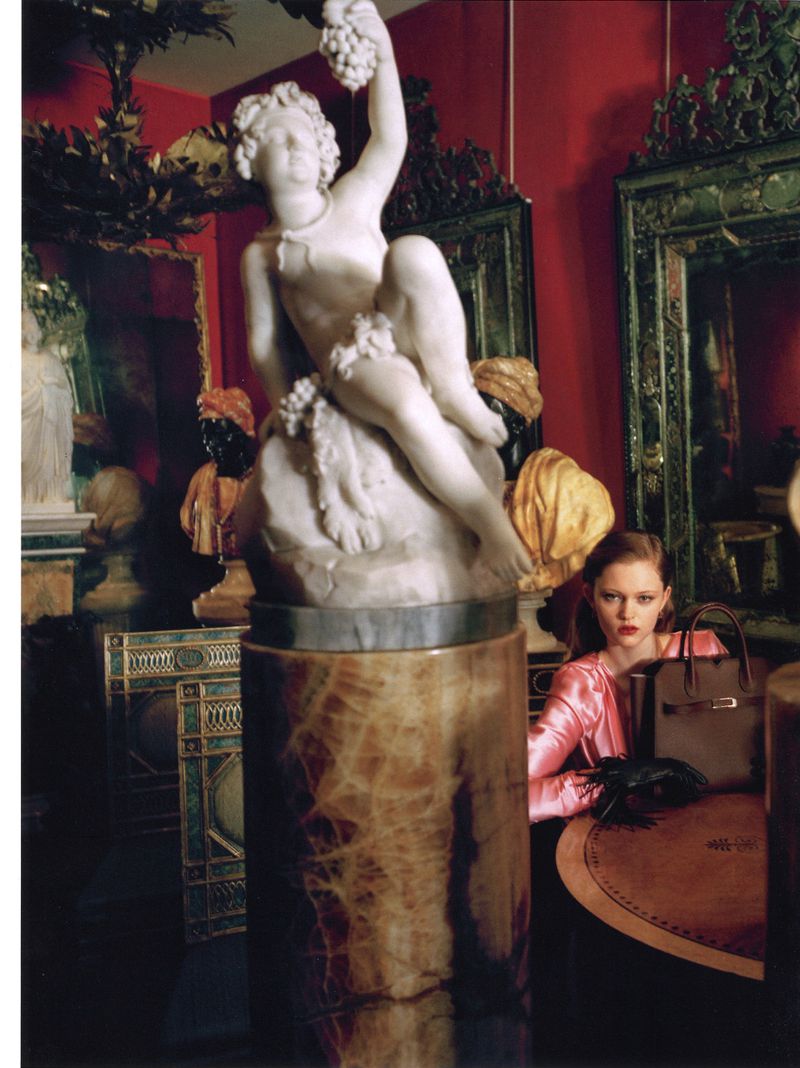 Indoor shot featuring a woman in a pink outfit sitting at a table with a handbag, set against a backdrop of a classical statue and red walls.