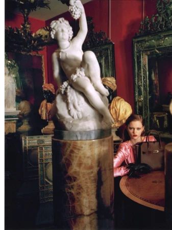 Indoor shot featuring a woman in a pink outfit sitting at a table with a handbag, set against a backdrop of a classical statue and red walls.