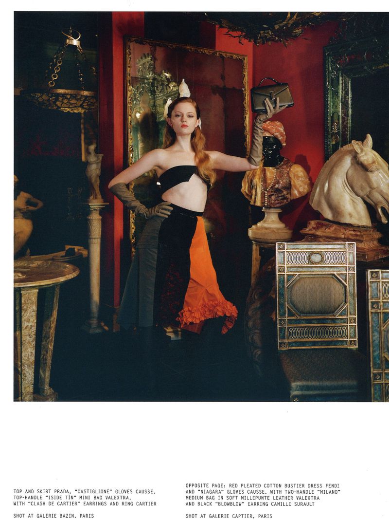 Fashion editorial shot of a model in an artistic indoor setting. The woman wears a black and orange top and skirt, gloves, and a headpiece.