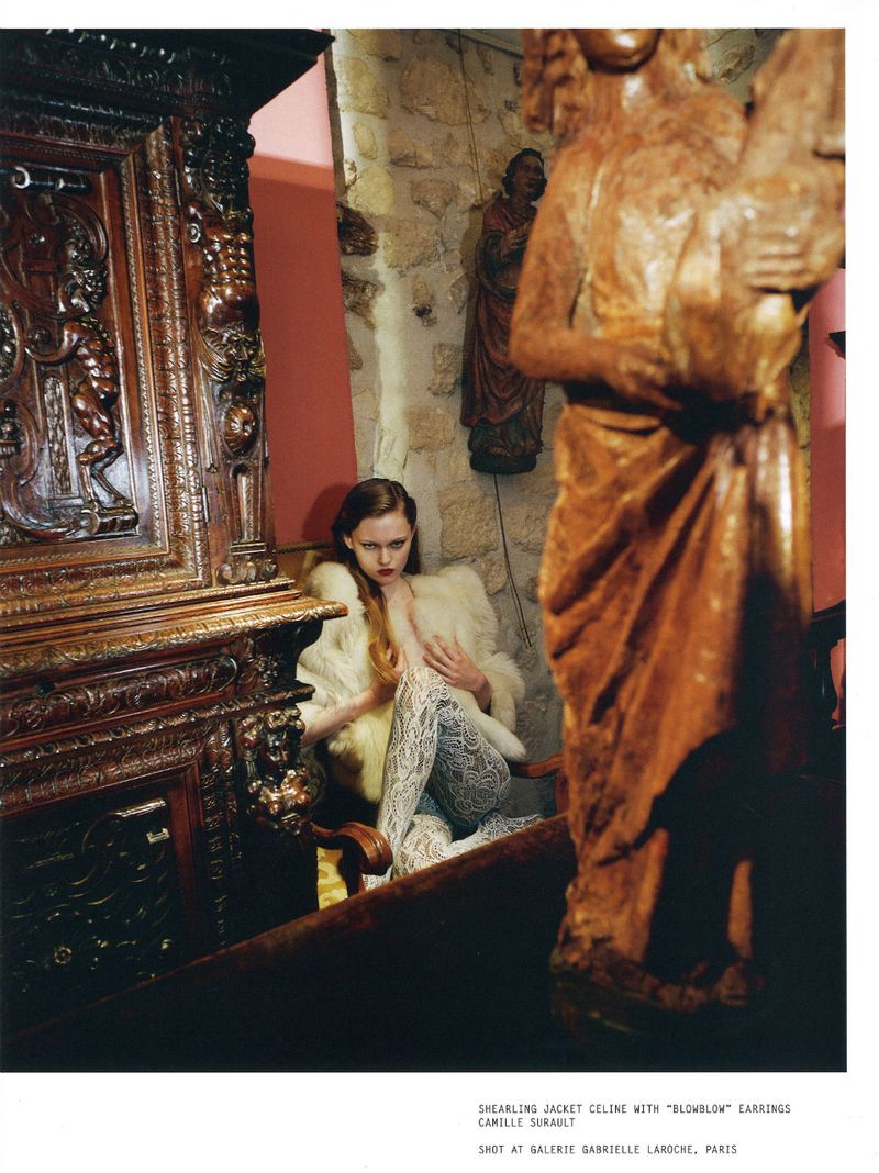 Fashion editorial featuring a woman in patterned leggings and a faux fur jacket, seated next to a large, ornate wooden sculpture.